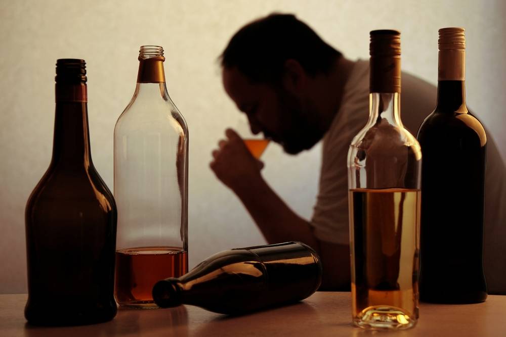 sombra de um homem bebendo de um copo com várias garrafas vazias a sua frente